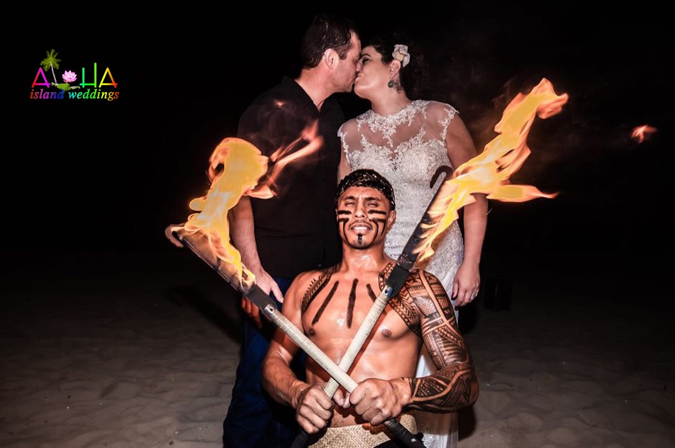 Bride and groom kiss behind a fire knife dancer holding two flaming knives at their night reception on the beach